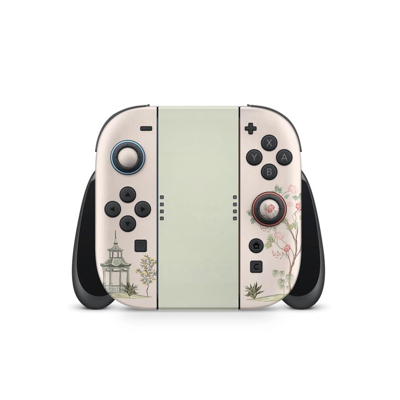 May include: A cream-colored gaming console with black handles and buttons. The left side features a small pagoda and trees, while the right side has floral designs. The center is a light green color.