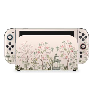 May include: A cream-colored Nintendo Switch console with a floral design featuring pink flowers, green trees, and a pagoda. The design covers the console's body and Joy-Con controllers, creating a cohesive aesthetic.