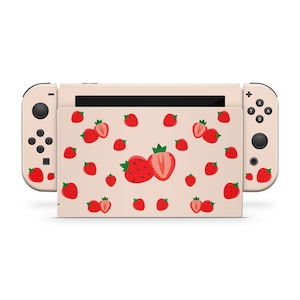 May include: A pink Nintendo Switch console with a custom skin featuring a repeating pattern of red strawberries on a light pink background. The console is closed and the Joy-Con controllers are attached.