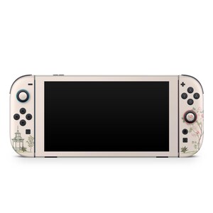 May include: A cream-colored gaming console with a black screen and floral design. The console features a pagoda and floral artwork on the sides. The buttons are black, and the joysticks are light blue and pink.