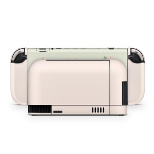 May include: A blush pink and white Nintendo Switch console. The console features a floral pattern on the top and sides. The design includes a light green background with pink flowers. The console has black accents.