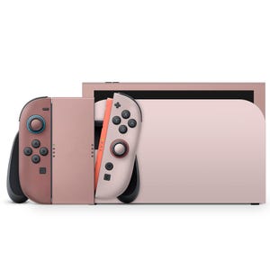 May include: A blush pink video game console with matching controllers. The console has a sleek design with a screen and a docking station. The controllers are detached and feature buttons and joysticks. The color scheme is a soft, monochromatic palette.