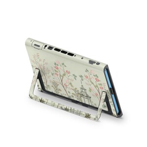 May include: A cream-colored electronic device with a floral design featuring pink blossoms and a pagoda. The device is propped up by a matching stand, showcasing the decorative pattern. The design wraps around the device and stand.