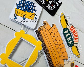 May include: A selection of party decorations themed around the TV show "Friends." The set includes a yellow picture frame, a Central Perk sign, a couch with the words "Pivot! Pivot! Pivot!", a film clapperboard with text, and a turkey wearing sunglasses.