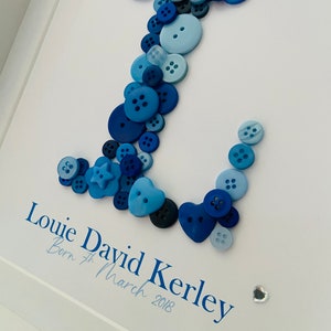 May include: A framed blue and white button art piece with the name "Louie David Kerley" and the date "Born 7th March 2018". The letter "L" is formed with blue buttons of various sizes and shapes.