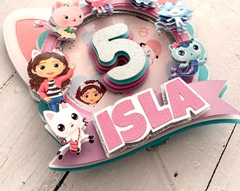 May include: A vibrant birthday cake topper with the name "ISLA" in pink letters with a silver glitter outline. The number "5" is central, surrounded by cartoon cat characters and a pink and teal colour scheme.