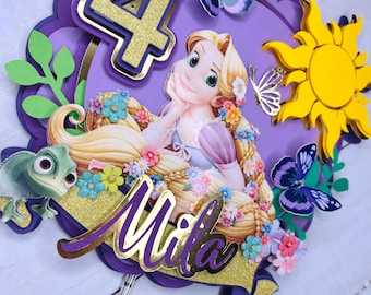May include: Purple, yellow and gold cake topper featuring Rapunzel from the Disney film Tangled. The topper includes a number 4, the name Mila, butterflies and a sun.