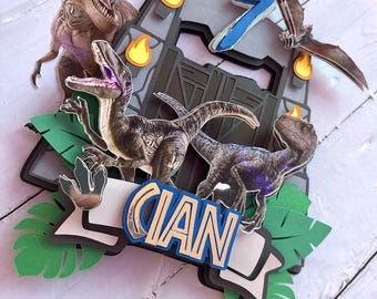 May include: A dinosaur-themed cake topper with a T-Rex, Velociraptors, and a Pterodactyl. The design includes a gate, flames, and the number 7. The name "CIAN" is displayed on a banner, with green leaves at the base.