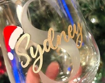 May include: A clear stemless wine glass with the name "Sydney" in gold script. A large frosted letter "S" is behind the name. A small Santa hat is on the left side of the glass. The background is a blurred Christmas tree.