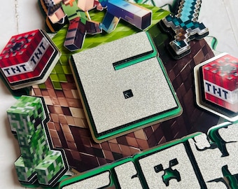 May include: A Minecraft-themed cake topper with a silver glitter finish. The topper features a green creeper, a green and black sword, a green and brown block with the number 6, and the word "CRAF" in green and black letters. There are also two red blocks with the word "TNT" on them.