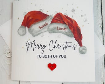 May include: A white Christmas card featuring two red Santa hats, each with a name. The card reads "Merry Christmas to Both of You" and has a small red heart. The design includes snowflakes and gold stars.