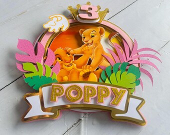 May include: A layered cake topper featuring two cartoon lion cubs, with the number 3 and the name "Poppy". The design includes pink and green tropical leaves, a gold crown, and a sunset background. The topper is designed for a birthday cake.