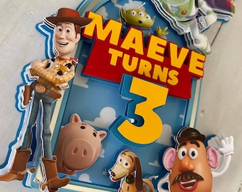 May include: A layered cake topper featuring characters from the Toy Story film. The topper includes Woody, Buzz Lightyear, an alien, Mr. Potato Head, Slinky Dog, and Hamm. The text "MAEVE TURNS 3" is displayed in yellow and red.