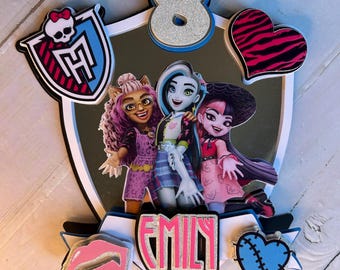 May include: A cake topper featuring three Monster High characters, a pink heart with a zebra print, a blue heart with stitches, a pink lip, a silver number 8, and the name "Emily" in silver glitter letters.