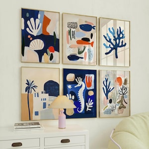 Può includere: Sei stampe artistiche astratte incorniciate con cornici dorate, raffiguranti la vita marina e scene costiere in tonalità di blu, arancione e bianco. Le opere d'arte sono esposte sopra un mobile bianco con una lampada e una pila di libri.