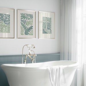 K&ouml;nnte beinhalten: Drei gerahmte botanische Kunstdrucke in Gr&uuml;n- und Blaut&ouml;nen h&auml;ngen &uuml;ber einer wei&szlig;en Badewanne. Die Drucke zeigen abstrakte Blatt- und Pflanzendesigns. Das Badezimmer hat eine klassische, spa-&auml;hnliche &Auml;sthetik mit einer wei&szlig;en Wanne und einer blauen Akzentwand.