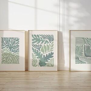 K&ouml;nnte beinhalten: Drei gerahmte Kunstdrucke mit botanischen Designs in Gr&uuml;n- und Blaut&ouml;nen. Die Drucke zeigen abstrakte Blatt- und Pflanzenformen auf cremefarbenem Hintergrund, eingefasst in goldfarbene Rahmen. Das Kunstwerk wird auf einem Holzboden vor einer wei&szlig;en Wand ausgestellt.
