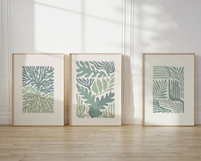 K&ouml;nnte beinhalten: Drei gerahmte Kunstdrucke mit botanischen Designs in Gr&uuml;n- und Blaut&ouml;nen. Die Drucke zeigen abstrakte Blatt- und Pflanzenformen auf cremefarbenem Hintergrund, eingefasst in goldfarbene Rahmen. Das Kunstwerk wird auf einem Holzboden vor einer wei&szlig;en Wand ausgestellt.