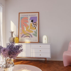 May include: A white dresser with three drawers and gold knobs. A white arch lamp with a gold accent sits on top of the dresser. A framed poster with the text "FLOWER MARKET" and abstract floral designs in pink, yellow, and blue is hanging on the wall behind the dresser. A vase with purple flowers sits on a pink round table in front of the dresser.