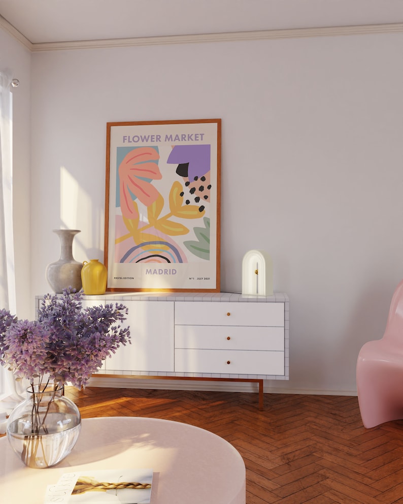 May include: A white dresser with three drawers and gold knobs. A white arch lamp with a gold accent sits on top of the dresser. A framed poster with the text "FLOWER MARKET" and abstract floral designs in pink, yellow, and blue is hanging on the wall behind the dresser. A vase with purple flowers sits on a pink round table in front of the dresser.