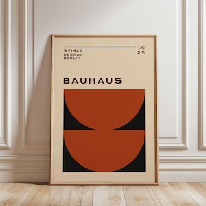 Puede incluir: Impresión artística Bauhaus enmarcada. La impresión presenta la palabra "BAUHAUS" en negro, con el texto "WEIMAR DESSAU BERLIN" encima. Debajo de la palabra "BAUHAUS" hay dos semicírculos en color naranja quemado, sobre un fondo negro. También se muestra el año "1923".