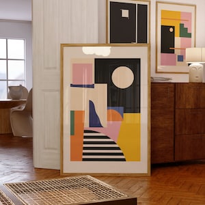 Puede incluir: Una impresión artística abstracta enmarcada con formas geométricas en negro, blanco, rosa, naranja y azul. La obra de arte incluye un gran círculo blanco y una forma de nube en la parte superior. Se ven otras dos impresiones enmarcadas en el fondo.