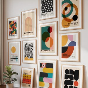 May include: A collection of framed Bauhaus art prints in various sizes and designs. The prints feature geometric shapes, bold colors, and the Bauhaus logo. The frames are light-colored wood, and the prints are displayed on a white wall.