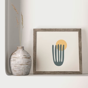 K&ouml;nnte beinhalten: Ein gerahmter Druck mit einem minimalistischen abstrakten Design, das eine dunkelgr&uuml;ne Form zeigt, die einer Pflanze &auml;hnelt, mit einem gelben Kreis dar&uuml;ber. Der Rahmen ist aus hellem braunem Holz.