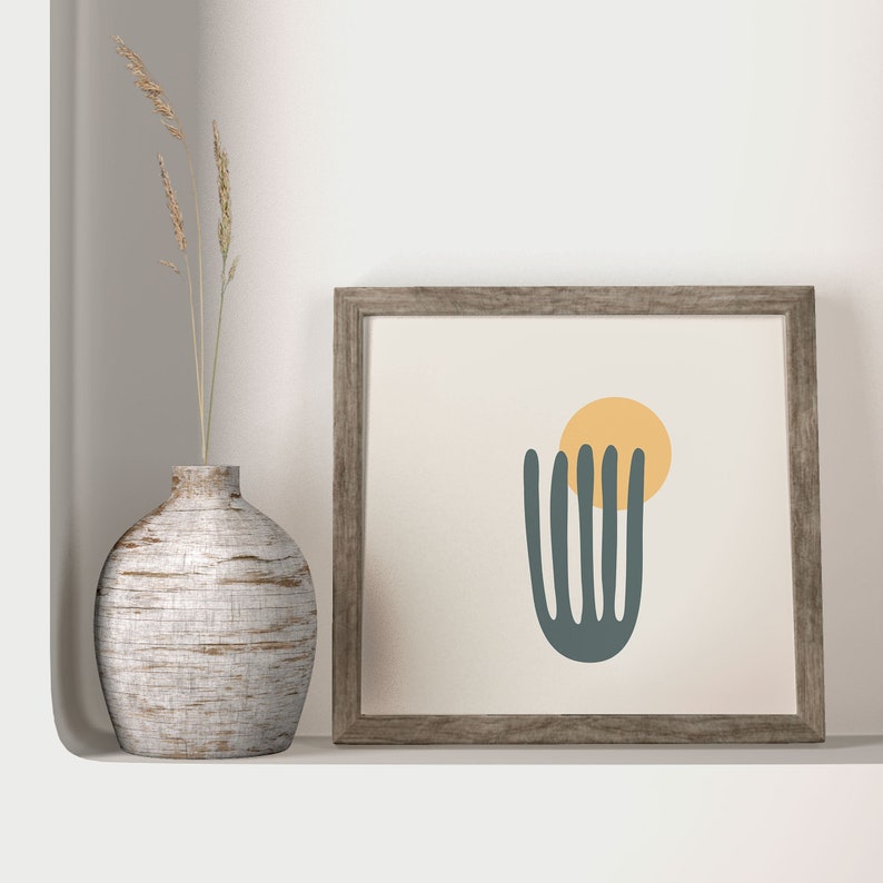 K&ouml;nnte beinhalten: Ein gerahmter Druck mit einem minimalistischen abstrakten Design, das eine dunkelgr&uuml;ne Form zeigt, die einer Pflanze &auml;hnelt, mit einem gelben Kreis dar&uuml;ber. Der Rahmen ist aus hellem braunem Holz.