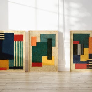 Può includere: Tre opere d'arte astratte incorniciate. Ogni pezzo presenta una cornice dorata e un design geometrico unico con blocchi di colore. I colori includono blu navy, rosso, verde, arancione, giallo e nero. L'arte è esposta contro un muro bianco.