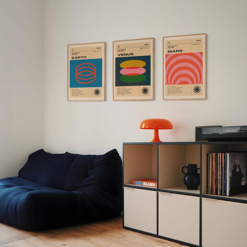 May include: Three framed posters with retro-style designs depicting Earth, Venus, and Mars. Each poster features a unique color palette and geometric shapes. A small orange lamp sits on a modern storage unit. A dark blue beanbag chair is in the foreground.