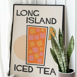 May include: A framed poster with a retro design featuring the text "Long Island Iced Tea" and a stylized illustration of a glass of iced tea. The poster is in a black frame and is leaning against a white wall.