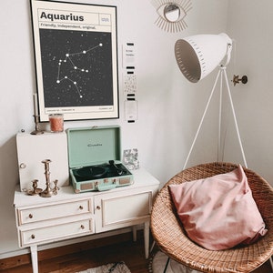 Puede incluir: Un tocadiscos blanco con un dise&ntilde;o de maleta verde se encuentra sobre una c&oacute;moda blanca con tres cajones. Un p&oacute;ster en blanco y negro con el texto "Aquarius, Friendly, Independent, Original" y un gr&aacute;fico de constelaci&oacute;n cuelga en la pared sobre la c&oacute;moda. Una almohada rosa se encuentra en una silla de mimbre con una alfombra blanca debajo.