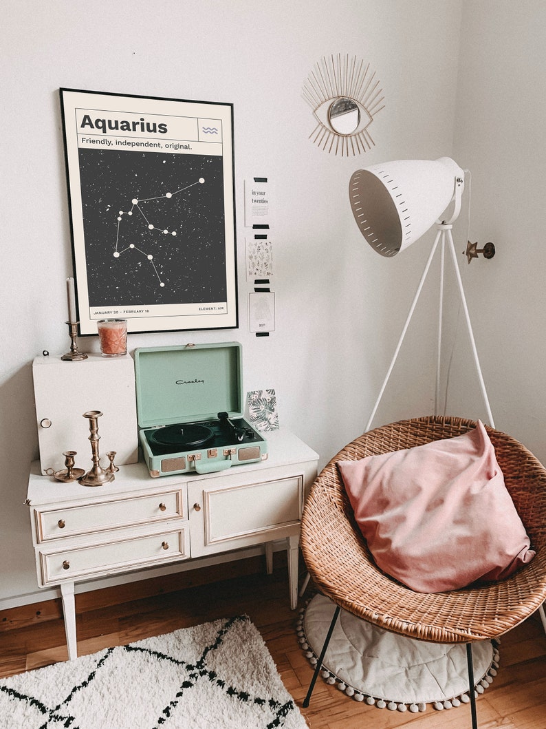 Puede incluir: Un tocadiscos blanco con un dise&ntilde;o de maleta verde se encuentra sobre una c&oacute;moda blanca con tres cajones. Un p&oacute;ster en blanco y negro con el texto "Aquarius, Friendly, Independent, Original" y un gr&aacute;fico de constelaci&oacute;n cuelga en la pared sobre la c&oacute;moda. Una almohada rosa se encuentra en una silla de mimbre con una alfombra blanca debajo.