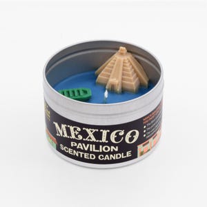 May include: A round tin candle with a blue wax base and a miniature replica of a Mayan pyramid. The candle is labeled "Mexico Pavilion Scented Candle".