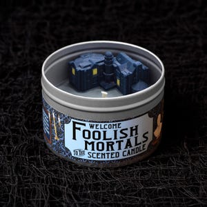 May include: A scented candle in a silver tin with a dark blue miniature haunted house on top. The label reads "Welcome Foolish Mortals to the Scented Candle" in black and white text with a blue floral pattern.