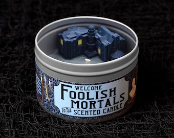 May include: A scented candle in a silver tin with a dark blue miniature haunted house on top. The label reads "Welcome Foolish Mortals to the Scented Candle" in black and white text with a blue floral pattern.