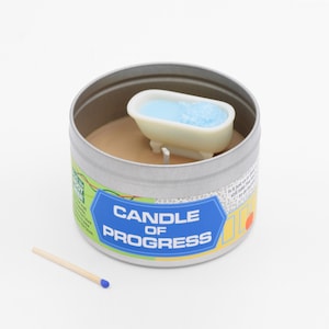 May include: A silver tin candle with a white bathtub figurine on top. The candle is labeled "Candle of Progress".