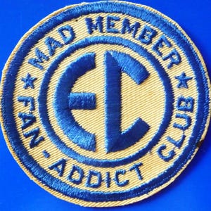 May include: A circular patch with blue embroidered text reading "MAD MEMBER CLUB FAN-ADDICT" on a tan fabric. A blue "EC" design is in the centre. The patch has a blue border, suggesting it's for a club or fan group.