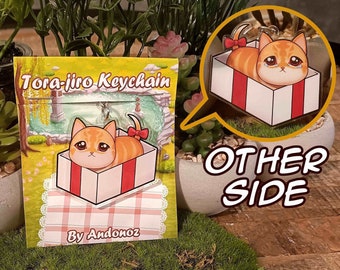May include: A keychain featuring a cartoon orange tabby cat inside a red and white gift box. The keychain is on a card that says "Tora-jiro Keychain" and "By Andonoz".