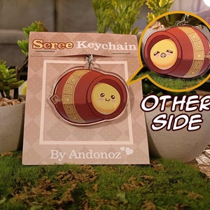 Könnte beinhalten: Ein roter und goldener Schlüsselanhänger mit einer Comicfigur darin. Der Schlüsselanhänger ist tonnenförmig mit einem niedlichen Gesicht und trägt die Aufschrift "Scree Keychain" und "By Andonoz".