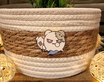 May include: A white and brown woven basket with a white and blue enamel pin of a cartoon ghost with stitches and a single eye. The pin is attached to the basket.