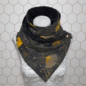 Peut inclure: Écharpe à motifs noirs et dorés avec fermeture boutonnée. Le motif répété présente des feuilles de ginkgo dorées et des formes géométriques sur fond sombre. L'écharpe est présentée sur une tête de mannequin blanche.