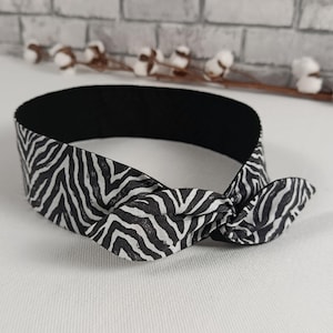Könnte beinhalten: Ein schwarz-weißes Zebra-Print-Haarband mit Schleife. Das Haarband hat eine schwarze Innenseite. Der Zebra-Print ist ein sich wiederholendes Muster aus schwarzen Streifen auf weißem Grund. Das Haarband ist etwa 2,5 cm breit.