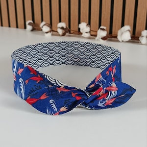 Könnte beinhalten: Ein Stoffstirnband mit blauem Hintergrund und einem Muster aus roten Koi-Fischen und weißen Wellenmotiven. Das Stirnband hat ein gedrehtes Knotendetail an der Vorderseite und ein kontrastierendes Innenfutter mit einem weißen und blauen Wellenmuster.