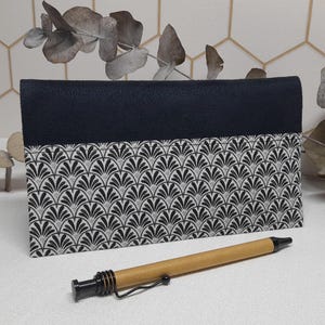 Peut inclure: Une pochette bleu marine avec un motif art déco noir et blanc. Un stylo marron avec une pointe noire est posé devant la pochette. La pochette est rectangulaire et texturée.