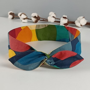Pode incluir: Uma faixa de tecido com um detalhe de nó torcido na frente. O acessório apresenta um padrão abstrato multicolorido e vibrante com formas arredondadas em tons de vermelho, amarelo, azul-petróleo, azul escuro e verde em toda a superfície.