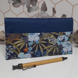 Peut inclure: Un portefeuille en tissu bleu avec un motif floral. Le portefeuille a un rabat en daim bleu foncé. Un stylo en bois est posé à côté du portefeuille.