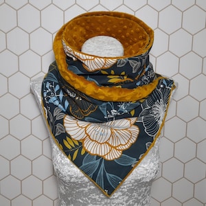 Puede incluir: Bufanda tubular floral en verde azulado oscuro y dorado. La bufanda presenta un gran diseño floral blanco con detalles dorados y azules. El interior es de tela dorada suave y texturizada. La bufanda se muestra en un maniquí.