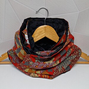 Pode incluir: Cachecol infinito vermelho e preto com padrão floral. O cachecol é feito de um tecido estampado com detalhes em vermelho, dourado e verde, e um forro preto de pelúcia. O cachecol é exibido em um cabide de madeira.
